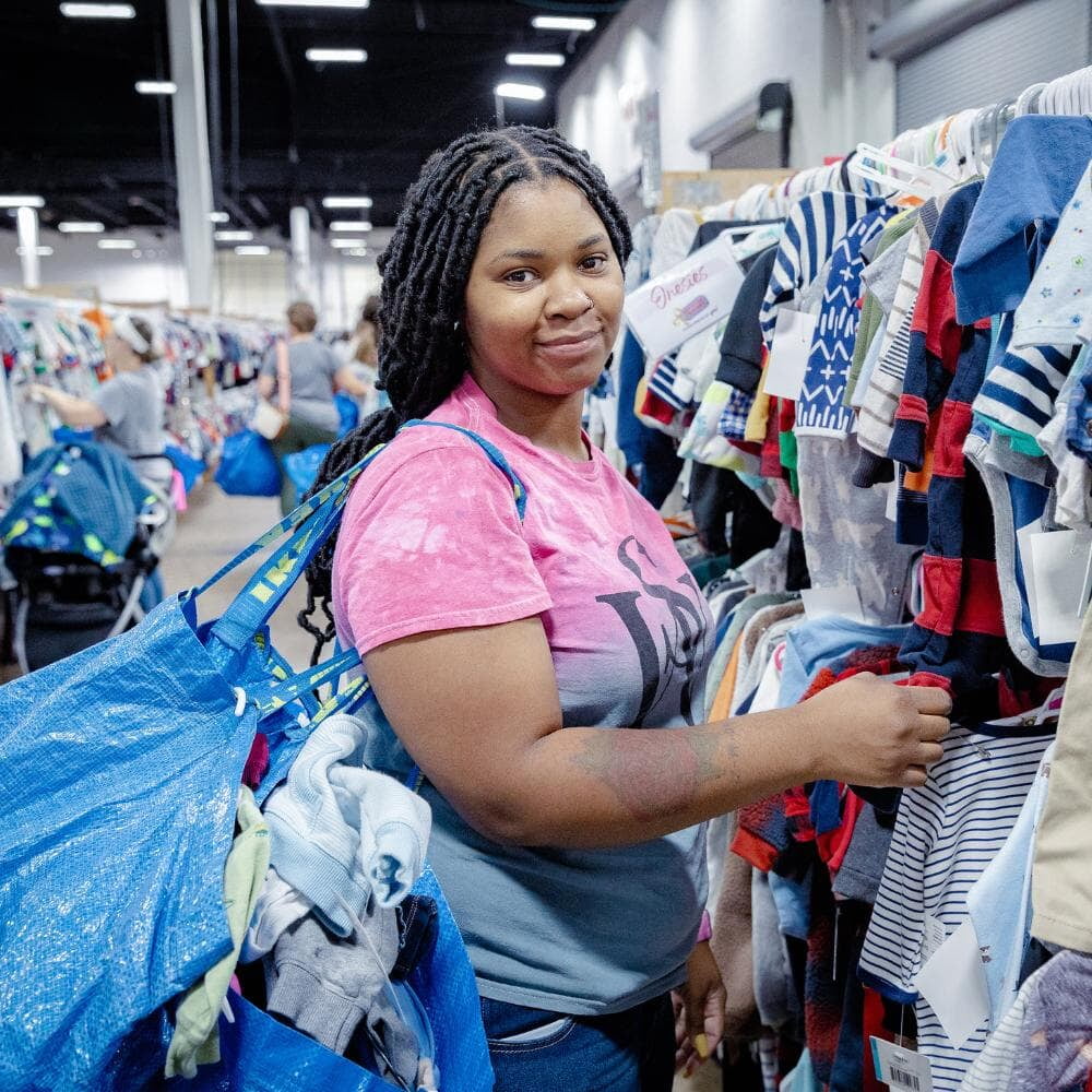 A mom with a large JBF shopping bag on her shoulder stands beside her husband who wears their toddler at a JBF sale.